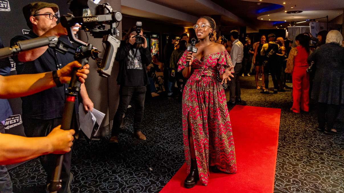 A red carpet host walking towards cameras holding a microphone wearing a long red dress.